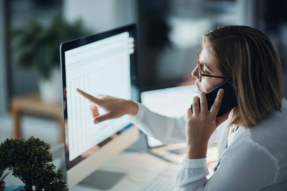 Kvinna sitter framför datorskärm och pratar i mobiltelefon