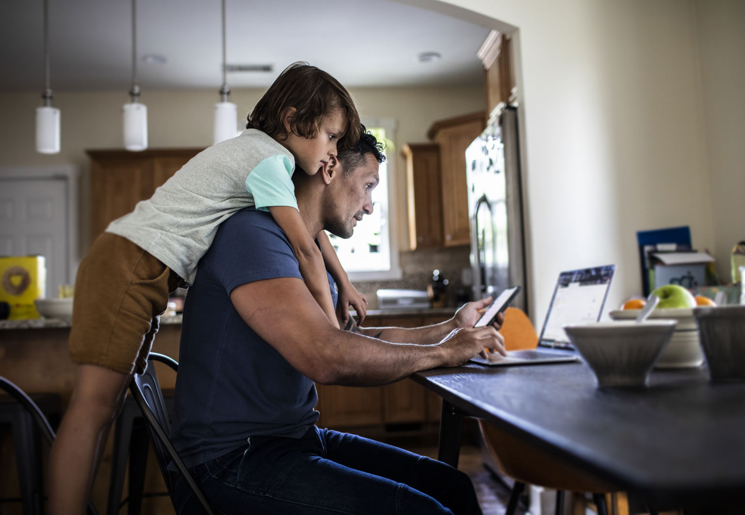 Pappa och son sitter vid matbordet och tittar ner i både telefon och dator och ser bekymrade ut