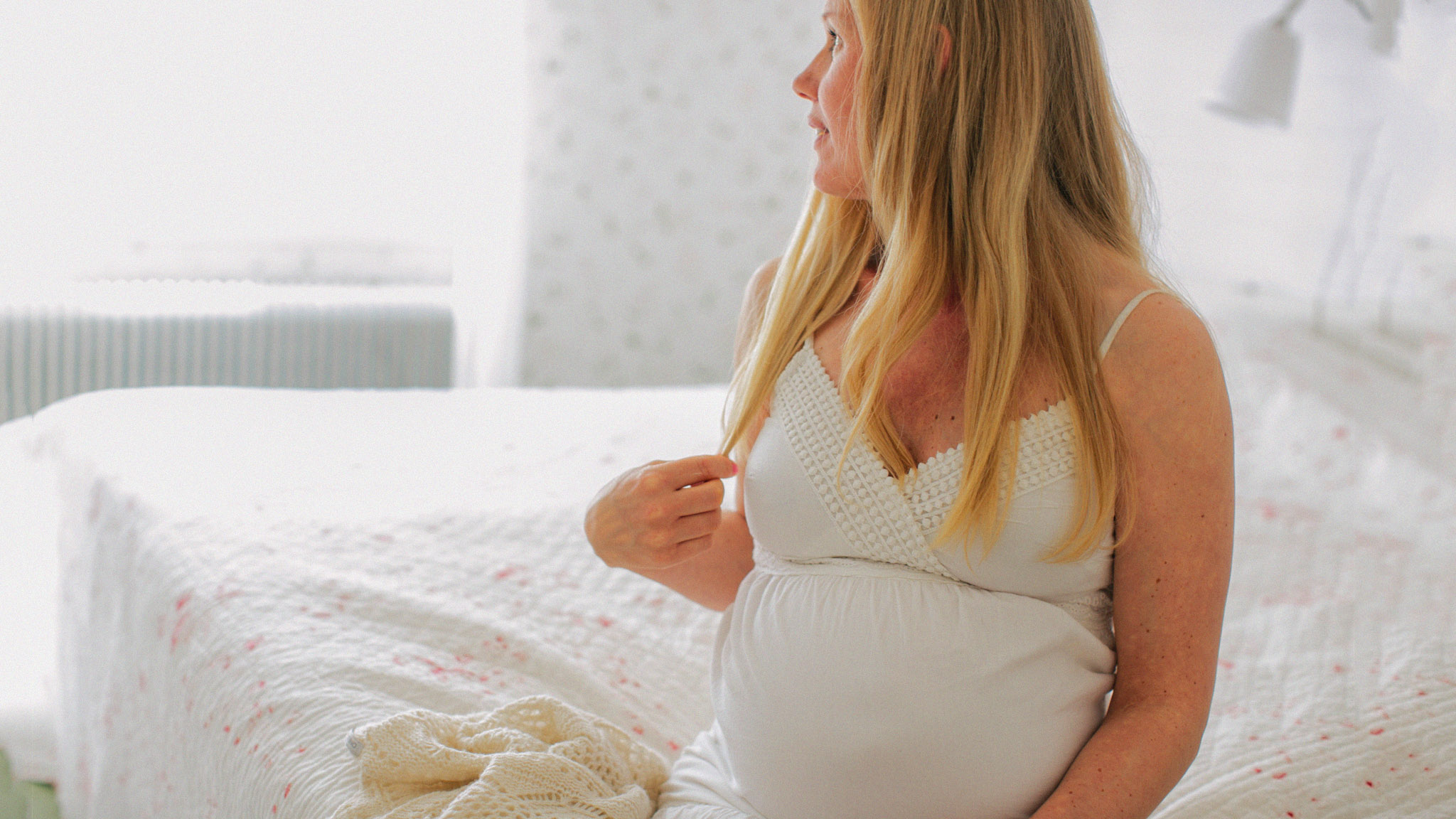 Höggravid kvinna sitter i sin säng och tittar ut genom fönstret