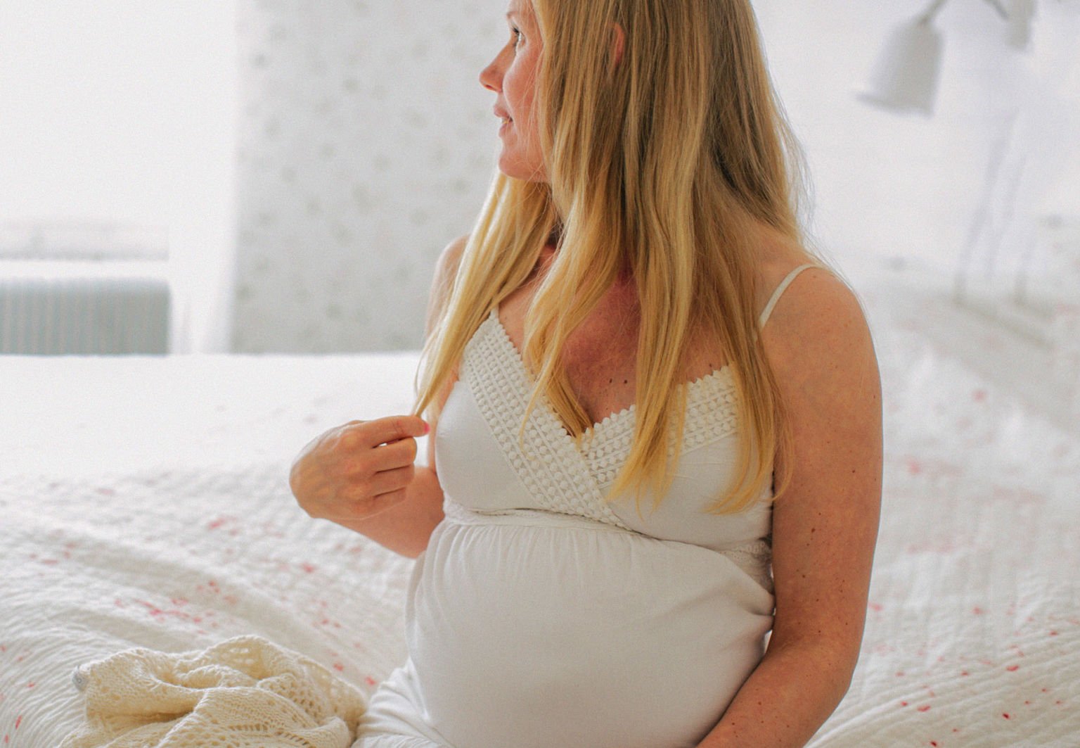 Höggravid kvinna sitter i sin säng och tittar ut genom fönstret