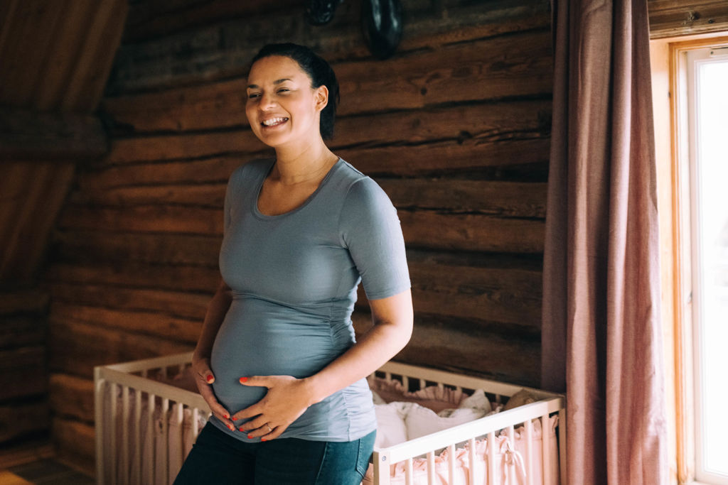 Kvinna står vid en spjälsäng och håller om sin gravida mage