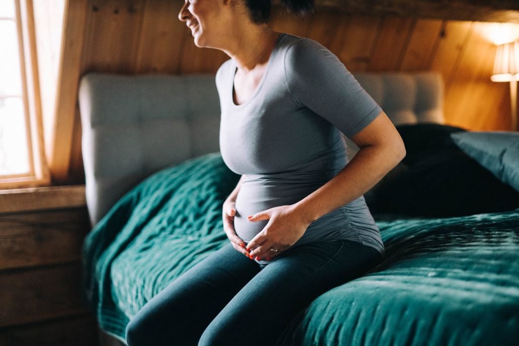 gravid kvinna i grå tröja sitter på sängkanten håller om sin mage