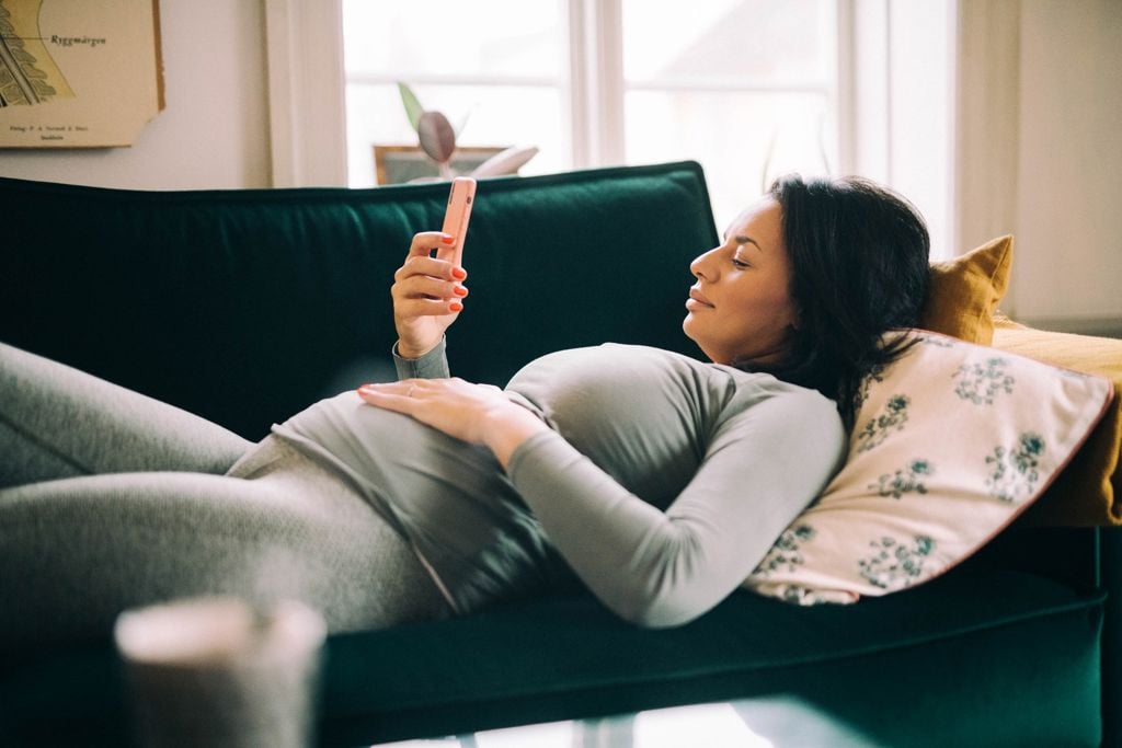 gravid kvinna i grå kläder ligger på en grön soffa och kollar på mobilen
