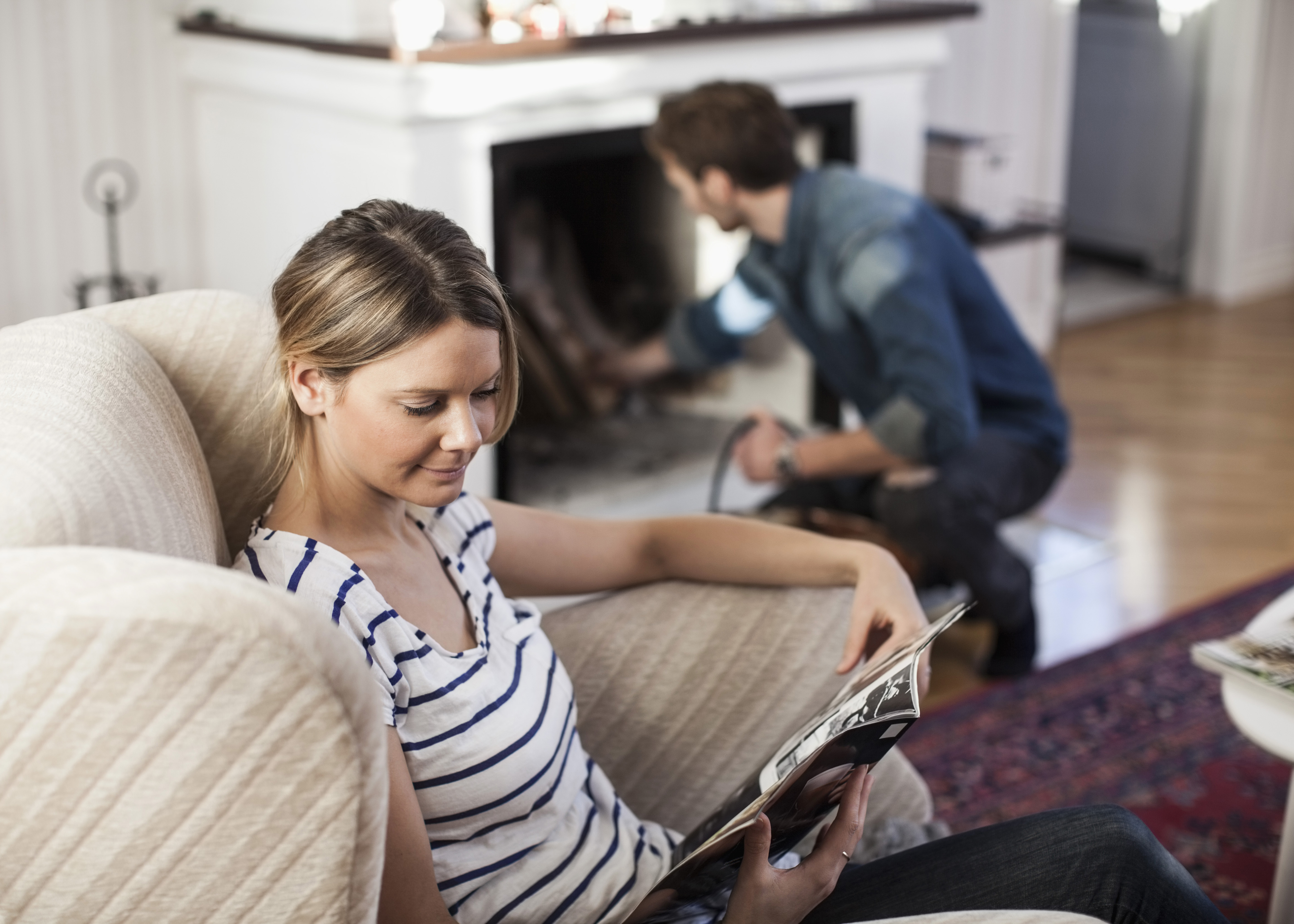 Kvinna läser en tidning i en fåtölj och en man tänder en brasa bakom henne