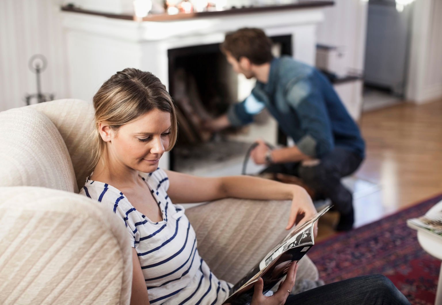 Kvinna läser en tidning i en fåtölj och en man tänder en brasa bakom henne