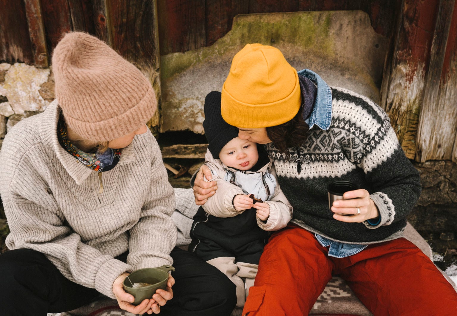 Litet barn i vinterkläder sitter mellan två vuxna som är varmt klädda och dricker något varmt utomhus i kyligt väder.