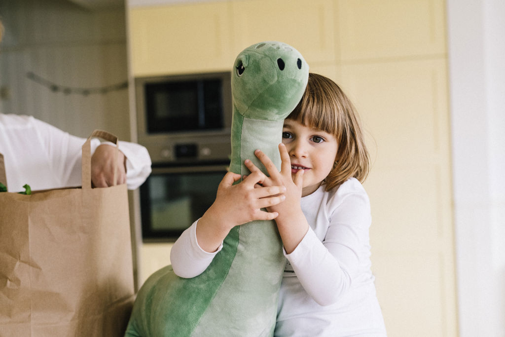 Barn kramar ett dinosauriegosedjur i köket brevid sin mamma som packar upp ur en matkasse