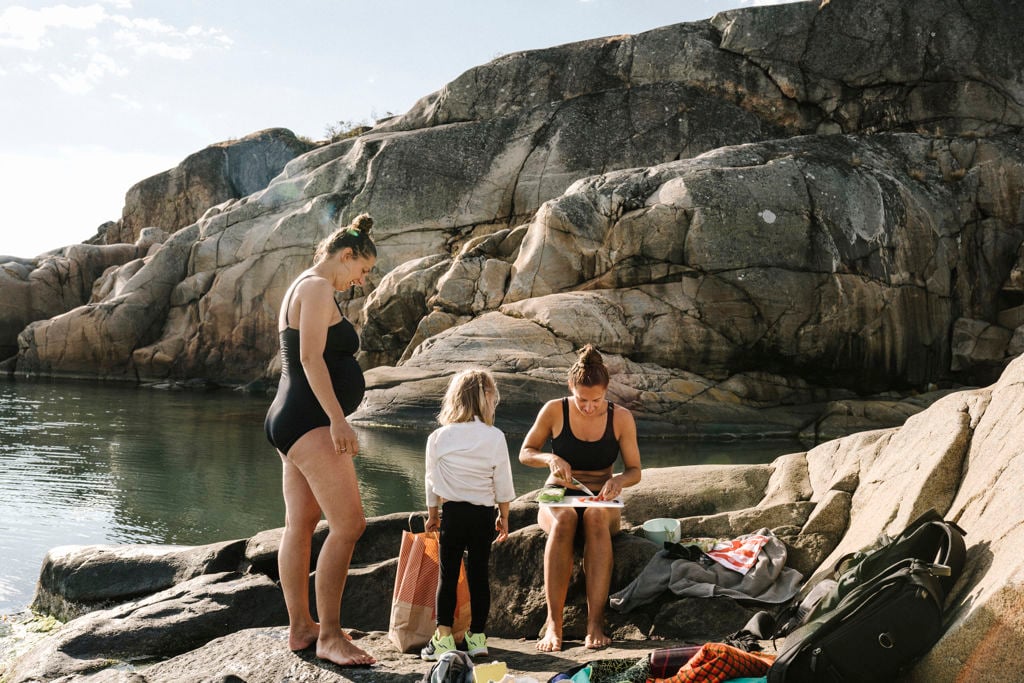 Gravid kvinna i baddräkt umgås med barn och annan vuxen vid vattnet vid klipporna
