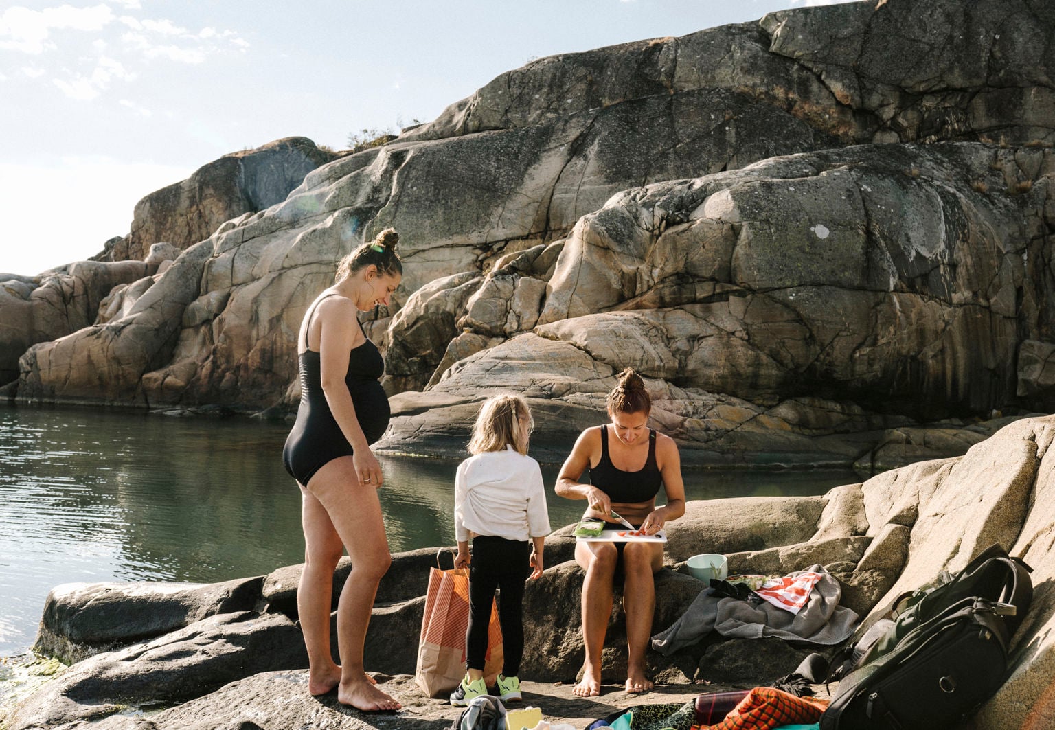 Gravid kvinna i baddräkt umgås med barn och annan vuxen vid vattnet vid klipporna
