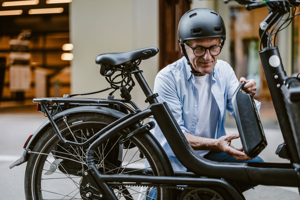 Äldre man med hjälm håller i elcykelns batteri på gatan