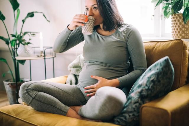 gravid kvinna i grå kläder sitter i soffan och dricker ur en mugg