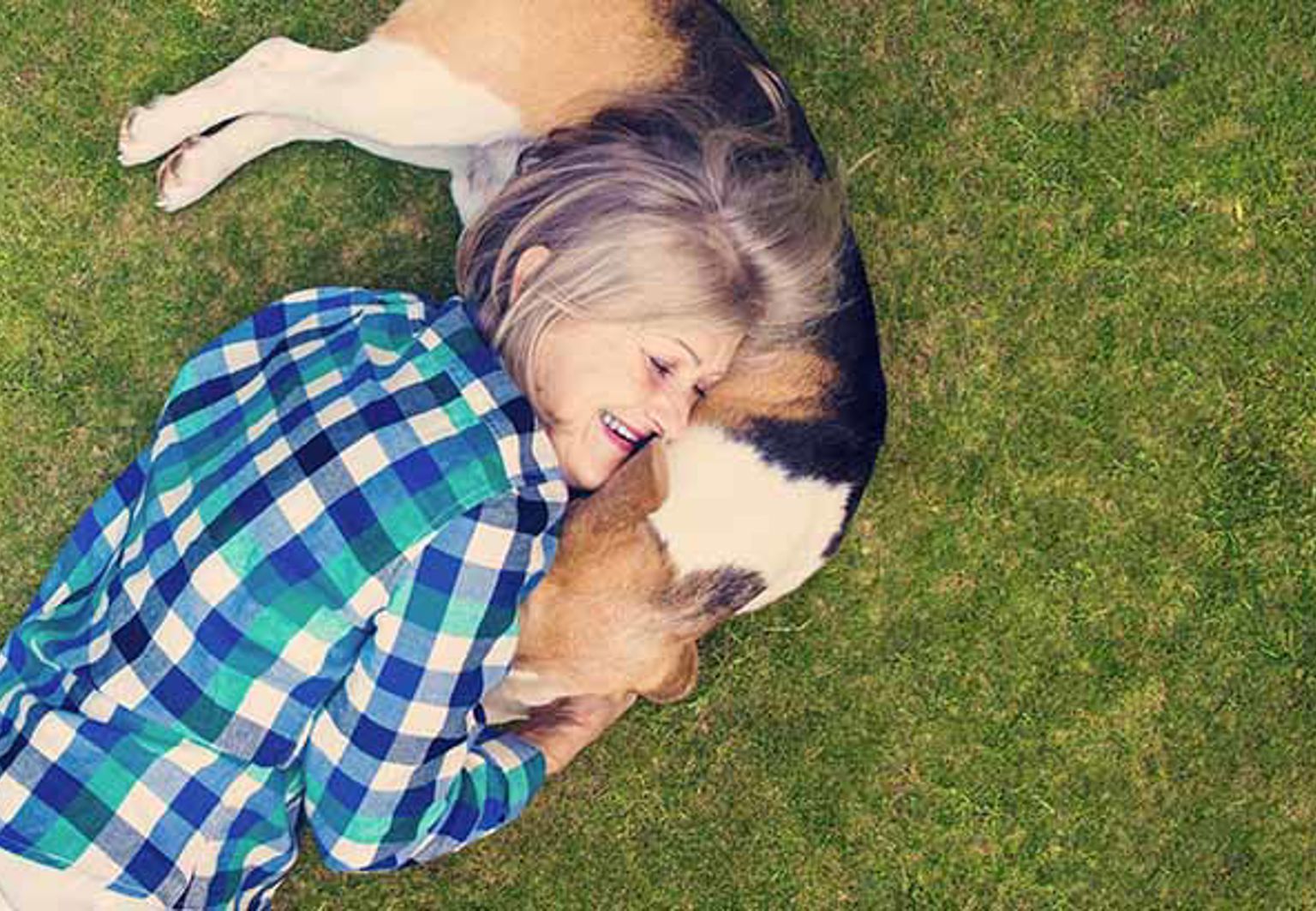 Kvinna och hund kramas liggandes på en gräsmatta