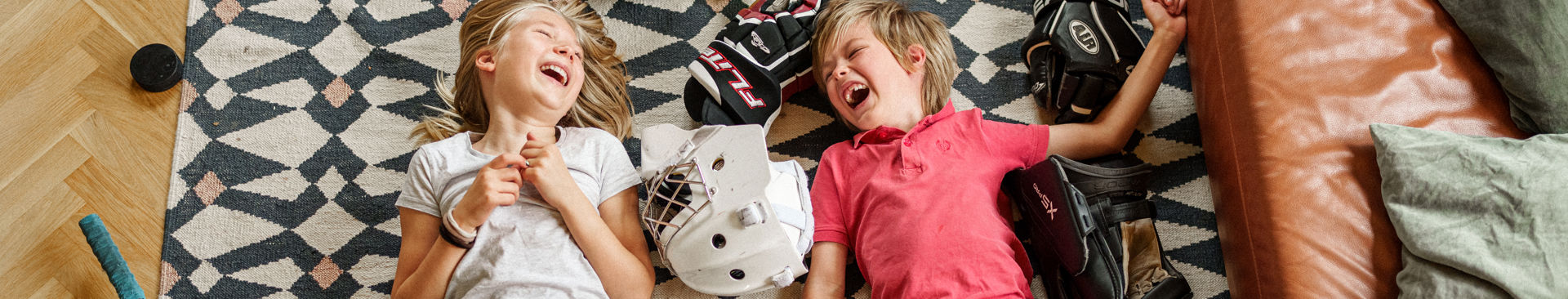 Två barn ligger på golvet och skrattar bland ishockey-utrustning